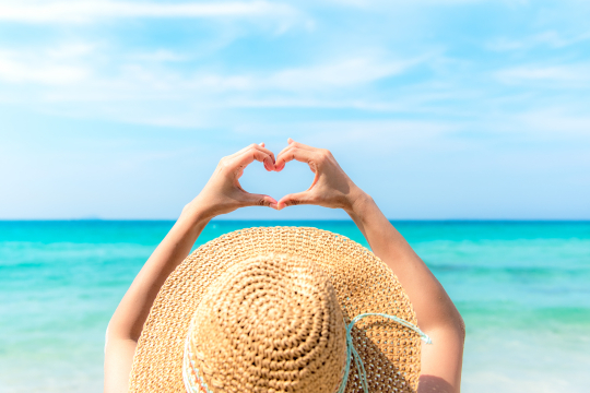 Frau mit großem Sonnenhut von hinten streckt ihre Hände nach oben und macht eine Herzform damit, im Hintergrund das Meer